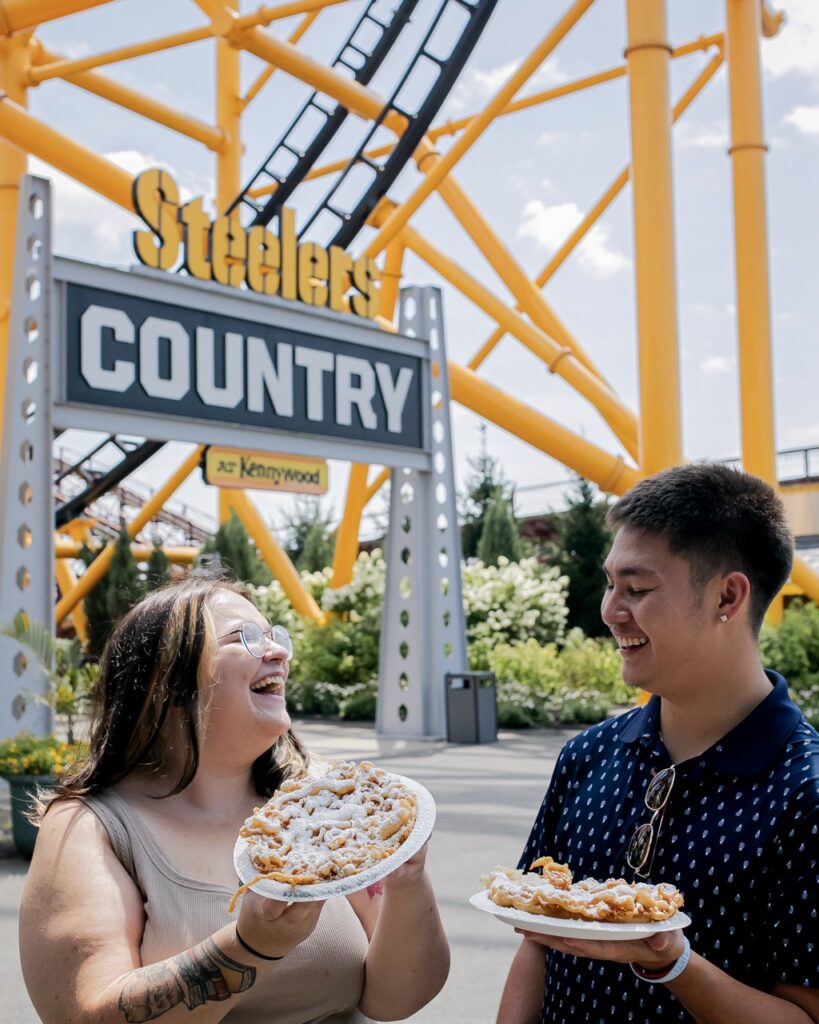 Two people hold funnel cakes in front of a roller coaster.