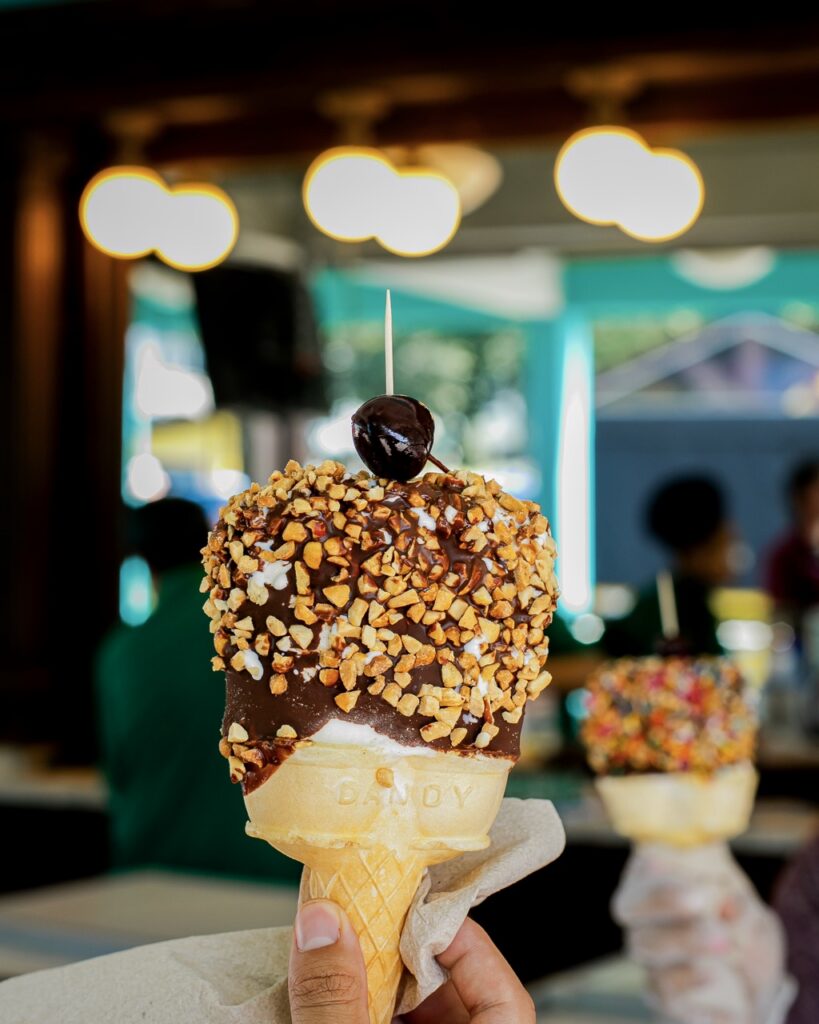 An ice cream cone with nuts on the chocolate and a cherry on top.