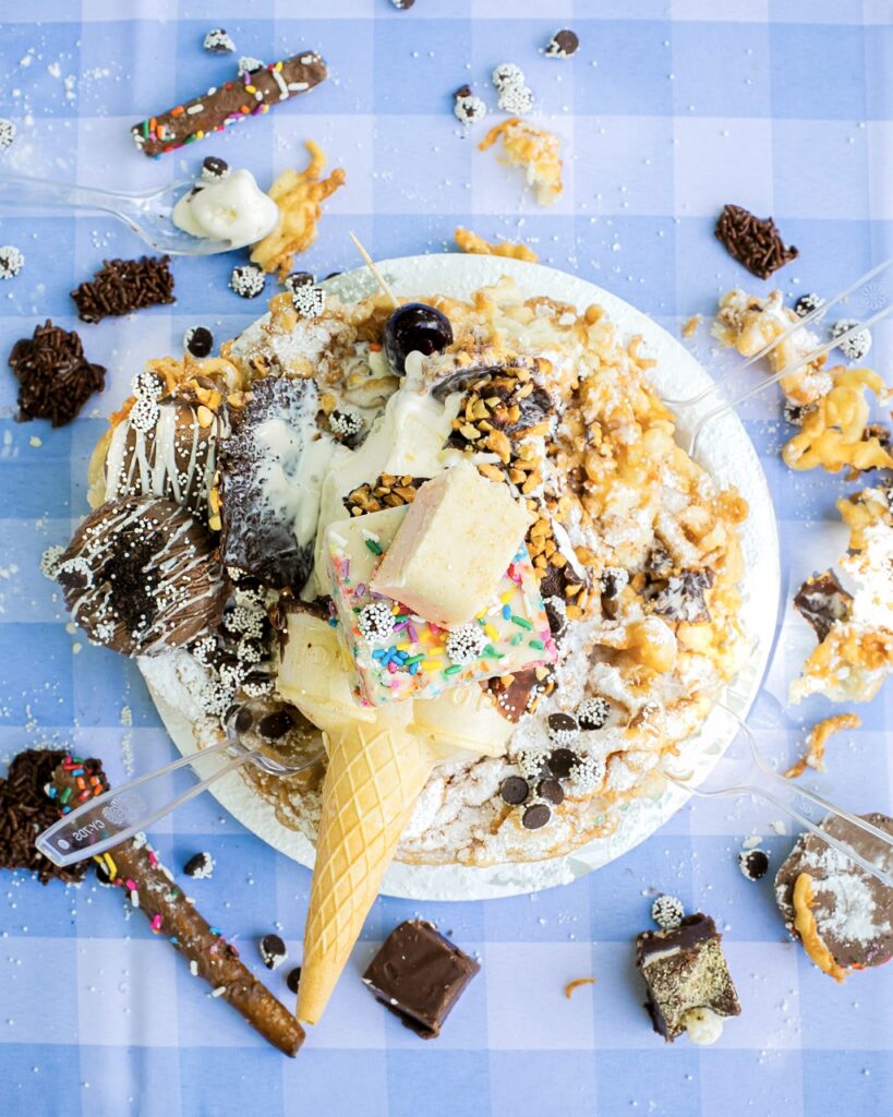 A funnel cake topped with ice cream, fudge, and other treats.
