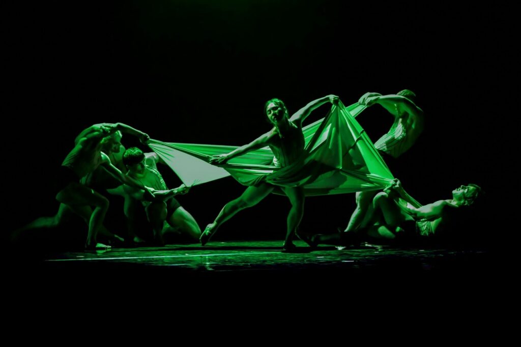 A group of people dance fluidly on stage in green lighting.