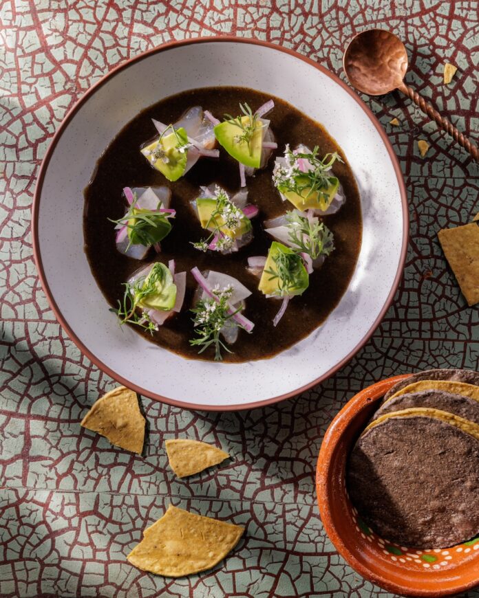 A bowl full of habanero aguachile with fluke fish and green garnishes beside a bowl of corn tortillas.