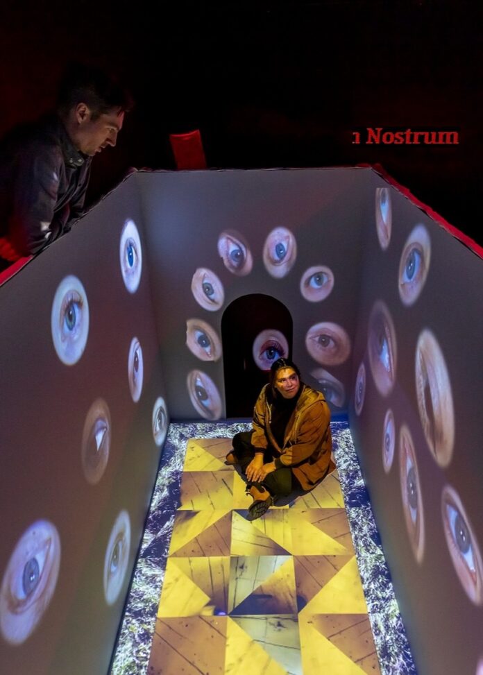 A person sits inside a box of projected eyeballs while another looks down at her from above at the Mattress Factory Pittsburgh Museum.
