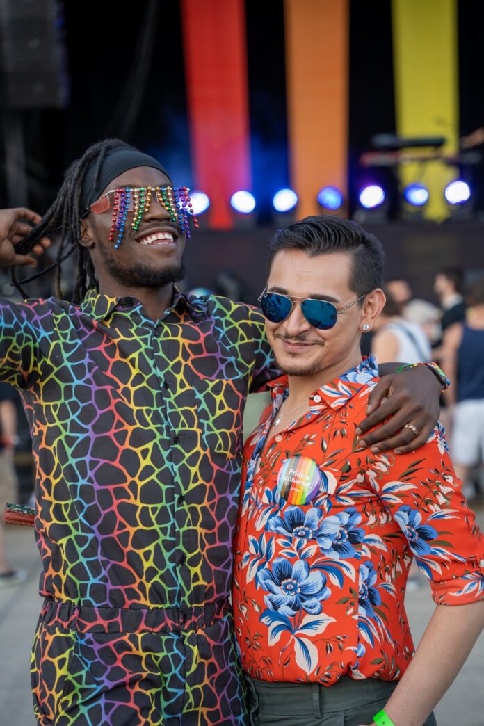 Two men in rainbow outfits and sunglasses stand in front of pride flag colors on a stage.