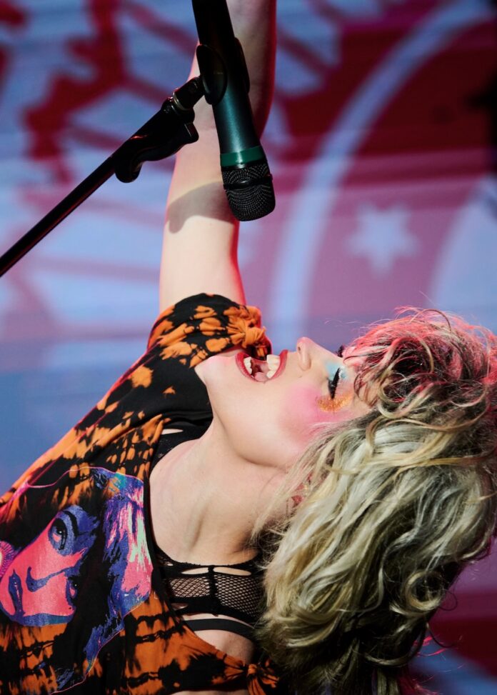 Hedwig looks up and sings into a microphone they're holding in the air.