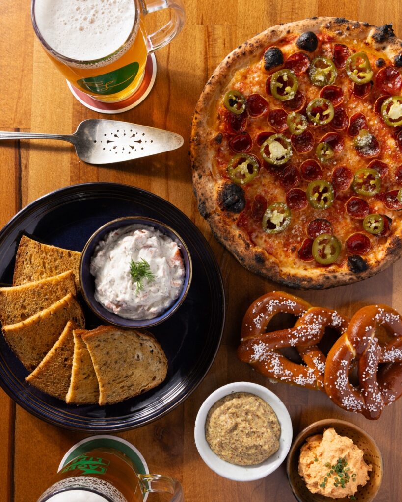 A round pizza with pepperoni and jalapeños sits on a wood table beside a plate of garlic bread and a white spread.