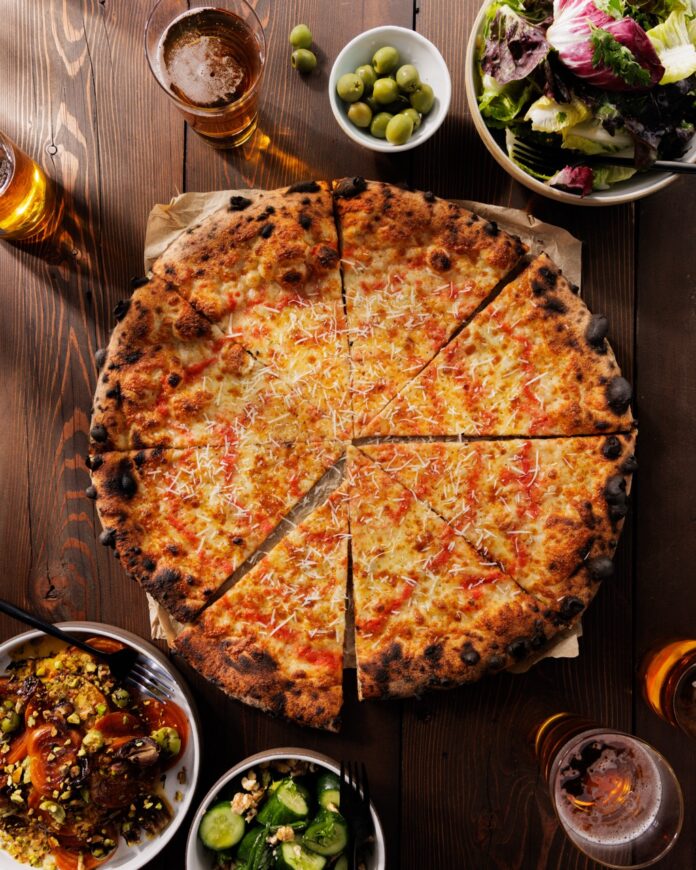 A large cheese pizza sits on a wood table with big and small bowls of sides and salads.