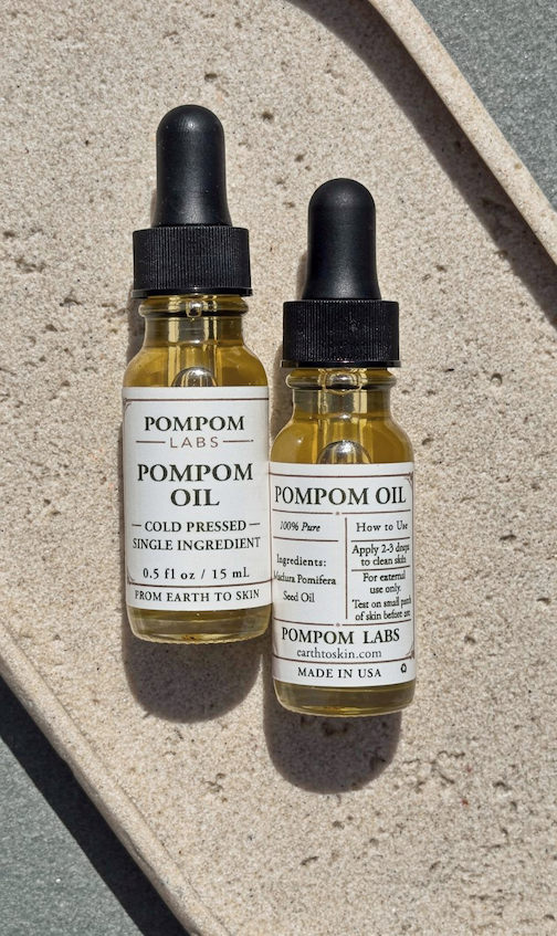 Two jars of Pom Pom Oil side-by-side with the front and back labels showing on a concrete surface.