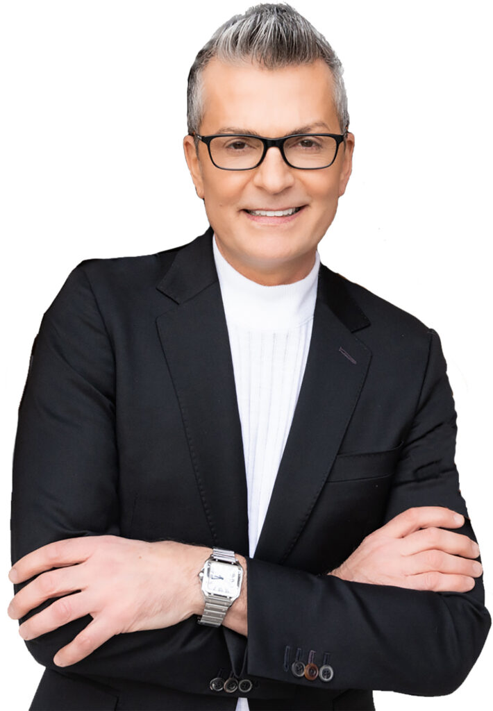 A photo of a man, Randy Fenoli, with his arms crossed wearing a white round collared shirt and a black blazer standing in from of a white background.