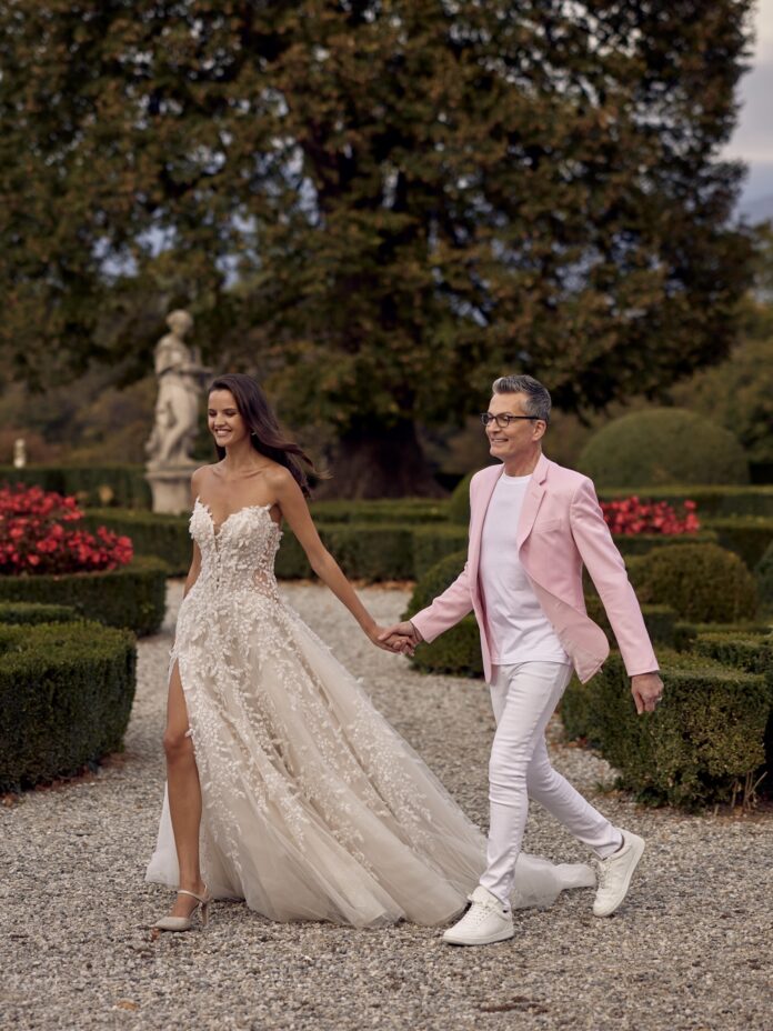 A picture of a man, Randy Fenoli, in a pink jacket walking through a courtyard hand-in-hand with a bride.