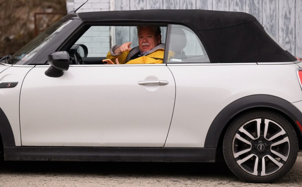 Rick Sebak in a yellow jacket points out the window of his white mini Cooper. 