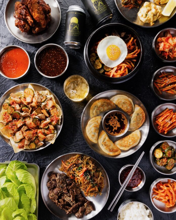 A black table full of plates featuring dumplings, Korean beef, stir fry, cans of beer, and little bowls of vegetable sides and sauces.