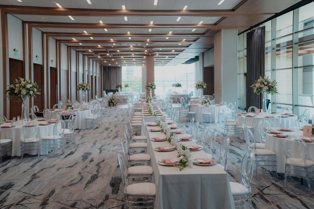 A photo of a ballroom during daylight hours set up for a wedding with a combination of long tables and round tables all decorated with white and pink.