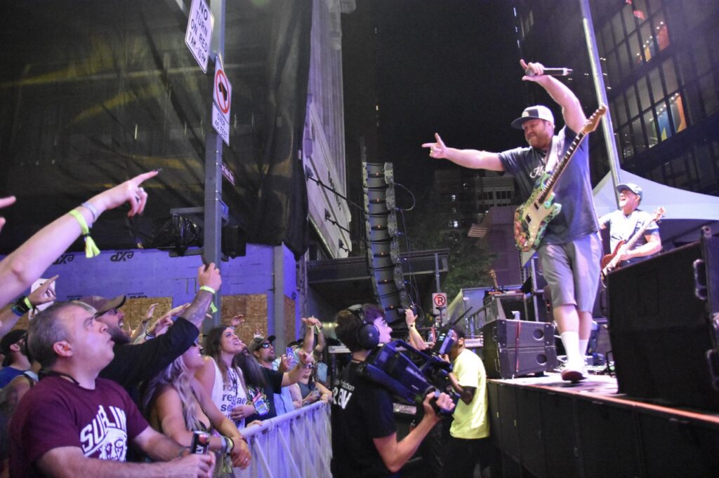 A man with a guitar points out to an audience behind a barricade fence.