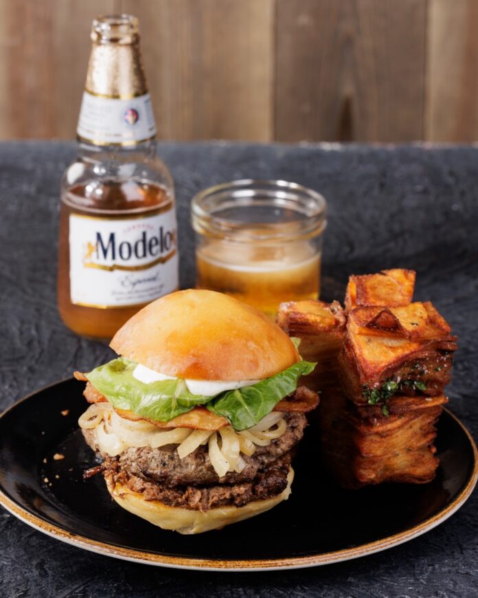 A burger with plenty of veggies on a balck plate with a stack of potatoes beside it and a beer behind it.