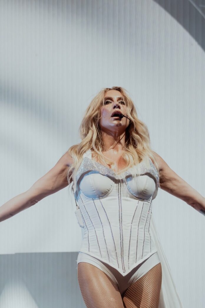Kesha stands in a white bodysuit corset in front of a white screen.