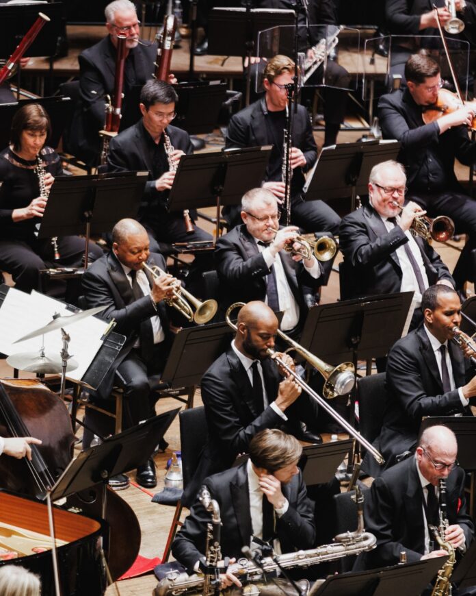 A section of a symphony with brass players and woodwinds behind them, sitting with music stands.