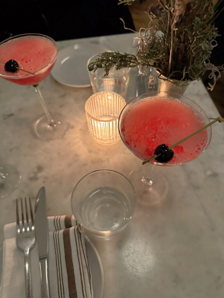 Two pink martinis with dark cherry garnishes on a white candlelit table. 