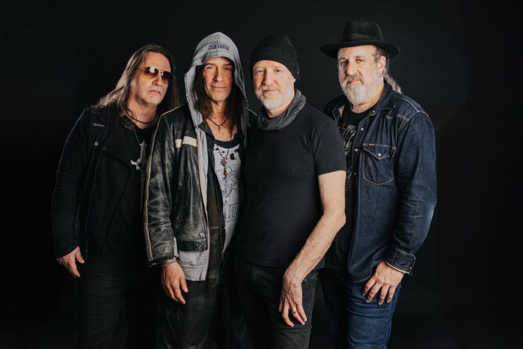 Four members of Spin Doctors stand together in front of a black background.