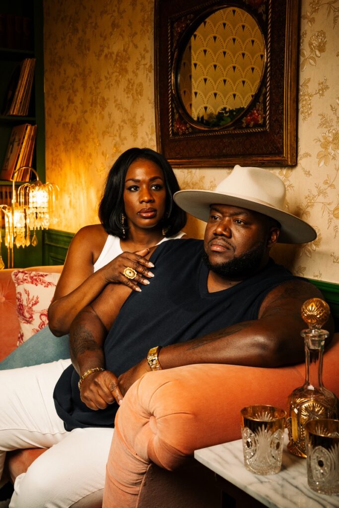 The two members of War and Treaty sit together on a brown couch, the man has a cowboy hat on and the woman has her hand on his shoulder.
