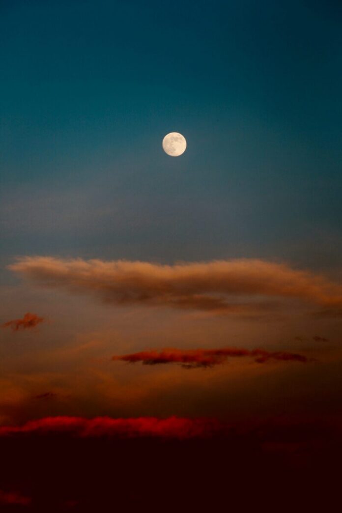 A full moon in a blue sky with orange-ish red clouds.