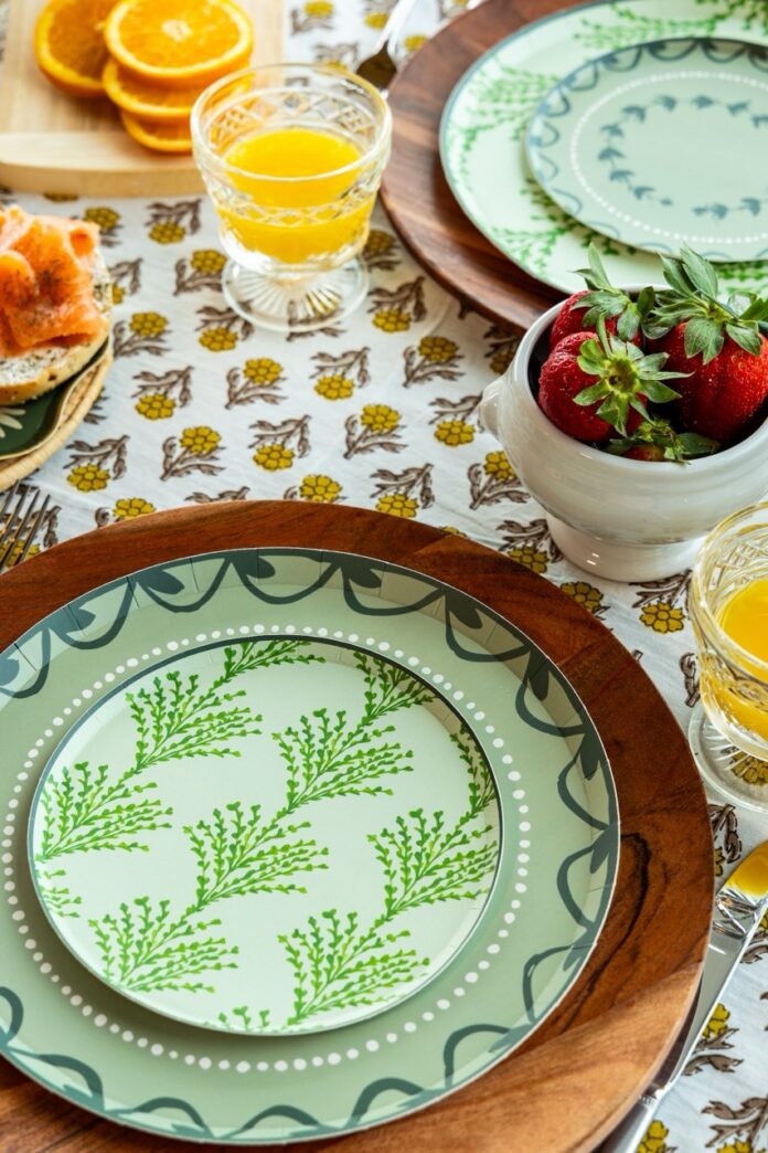 A stack of green paper plates on a pattern tablecloth with a glass of orange juice above it.