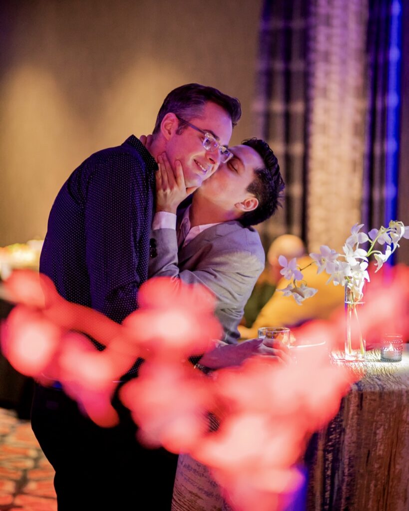 One man kisses another's cheek.
