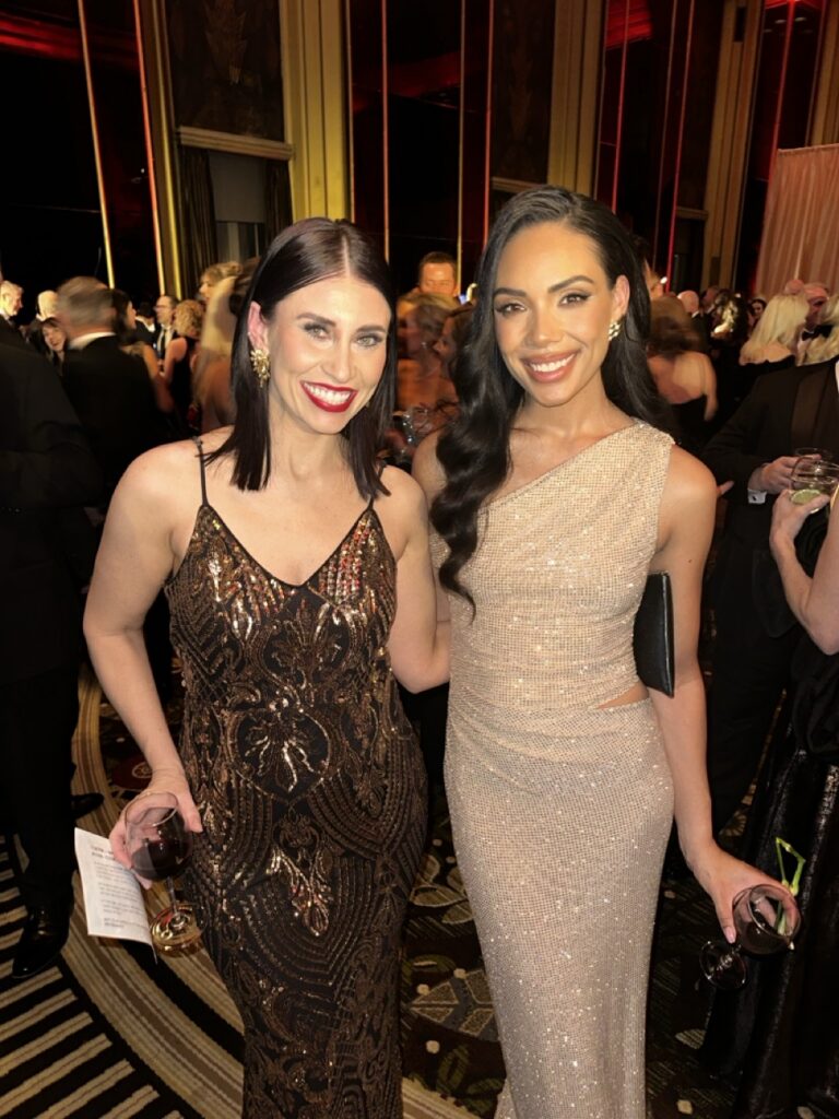 Two women in long sparkly dresses stand together.