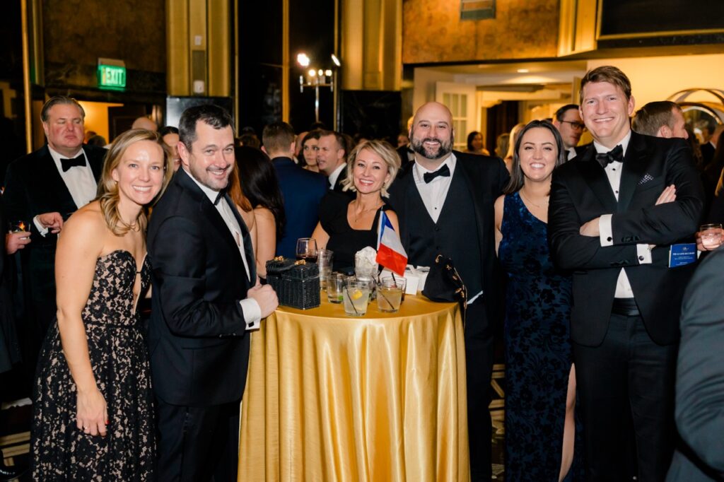 A group of people in evening-wear stand around a tall table with a gold table cloth and French flag.
