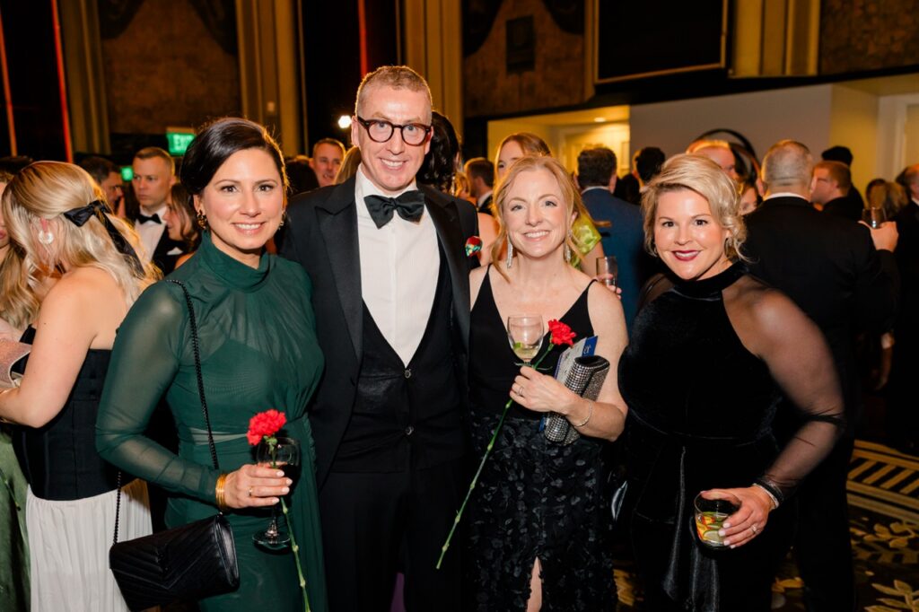 Three women and a man in a suit stand together, red carnations in hand.