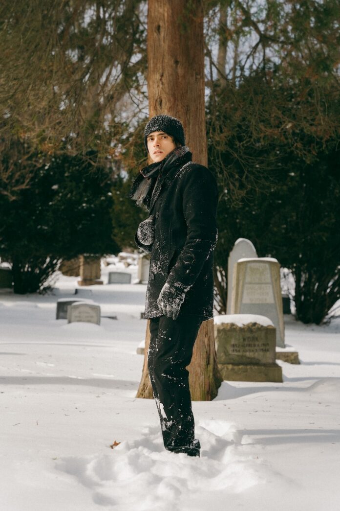 A man walks through snow in a winter jacket and hat and gloves in a graveyard.