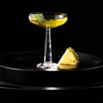 A tall coupe glass with a yellow sage-infused gin cocktail and a slice of pineapple beside the glass.