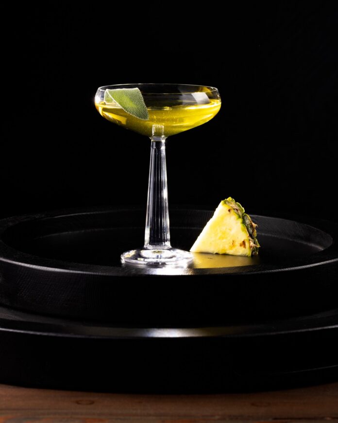 A tall coupe glass with a yellow sage-infused gin cocktail and a slice of pineapple beside the glass.