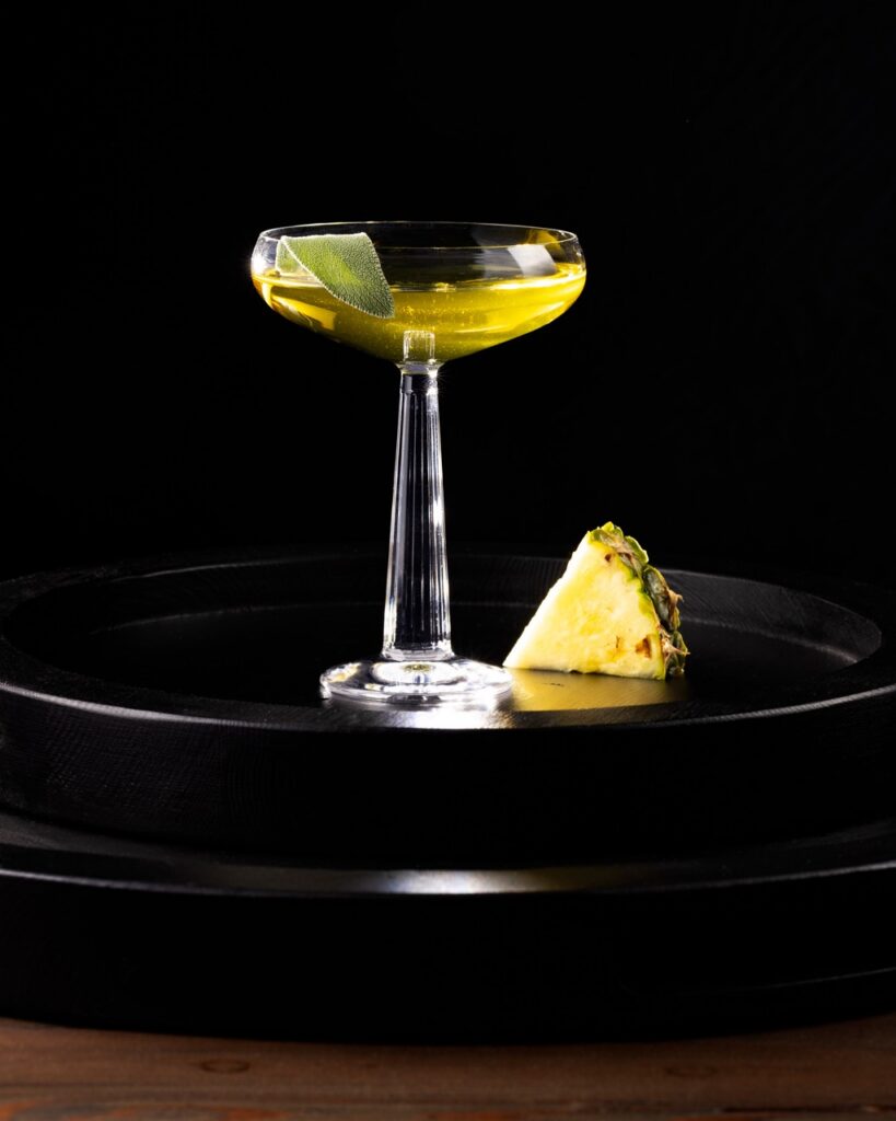 A tall coupe glass with a yellow sage-infused gin cocktail and a slice of pineapple beside the glass.