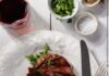 A Carnivore’s Guide to the Best Steaks, BBQ, and Beyond in Pittsburgh A white plate with a cut up NY strip steal on it with silverware beside the meat, seasonings in the right hand corner of the table, and a red glass of wine on the elft side of the plate.
