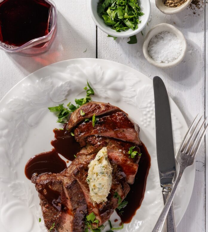 Best Steaks BBQ Meat Pittsburgh TABLE Magazine A white plate with a cut up NY strip steal on it with silverware beside the meat, seasonings in the right hand corner of the table, and a red glass of wine on the elft side of the plate.