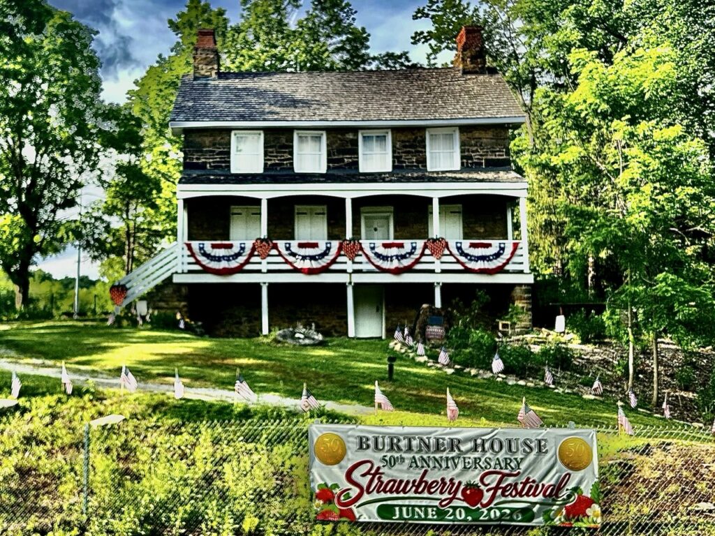Burtner House in the green grass with trees around it and a banner for the Strawberry Festival.
