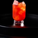 A PB&J cocktail in red and orange tones sits in a glass with ice on a black tray.
