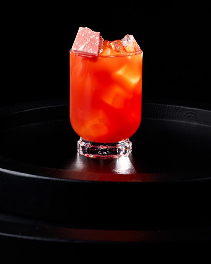A PB&J cocktail in red and orange tones sits in a glass with ice on a black tray.