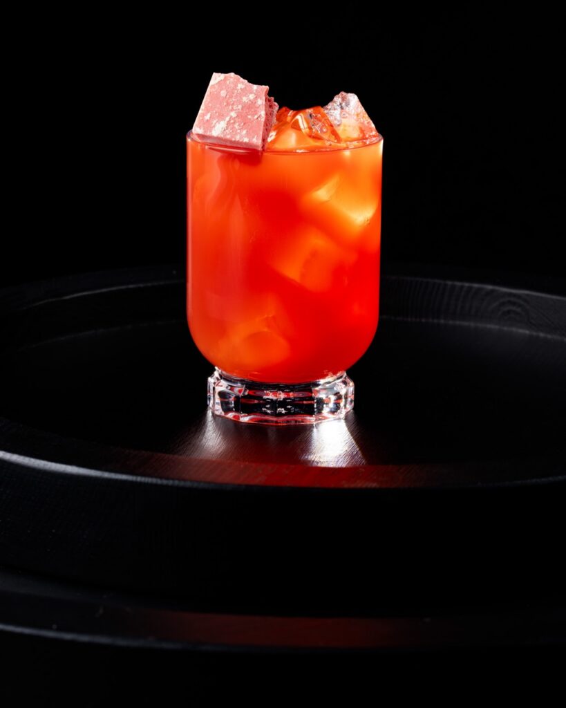 A PB&J cocktail in red and orange tones sits in a glass with ice on a black tray.