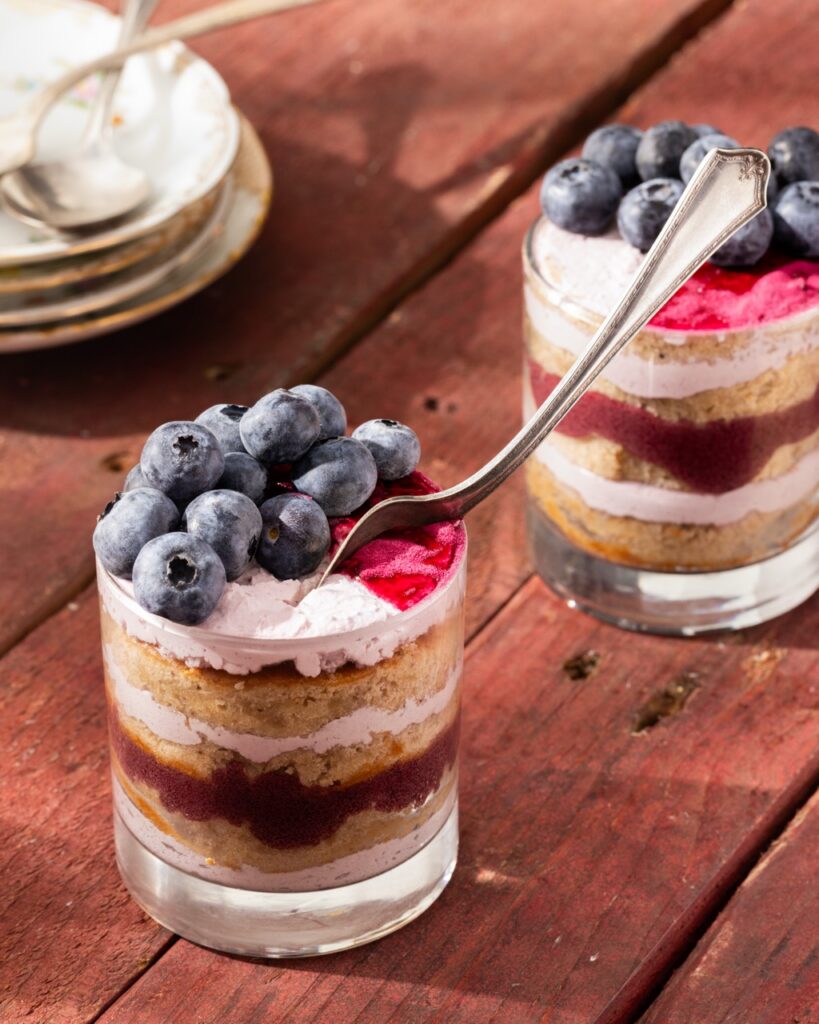 A spoon sticks out of a chai tea cake cup with blueberries on top in front of another chai tea cake cup with blueberries on top.