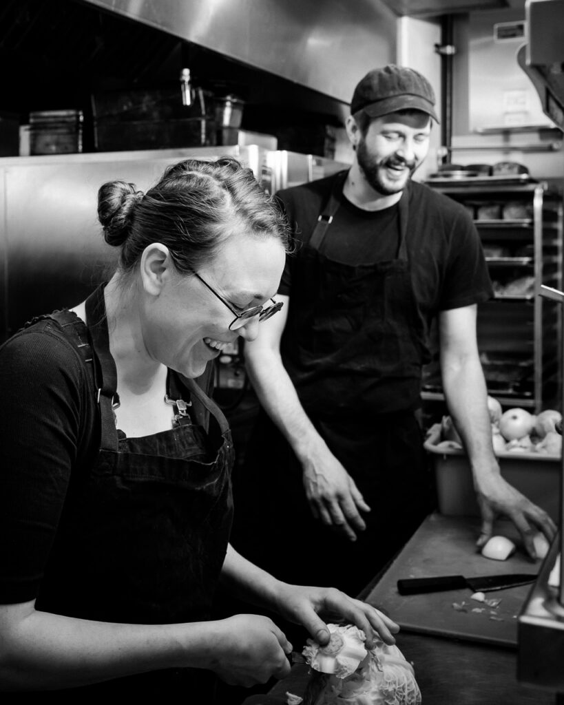 Chefs Tomasz and Kate from Apteka in black aprons as Tomasz wears a hat and they work at the counter.