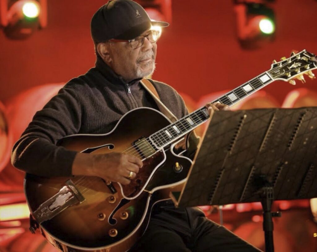 A man plays guitar sitting behind a music stand.