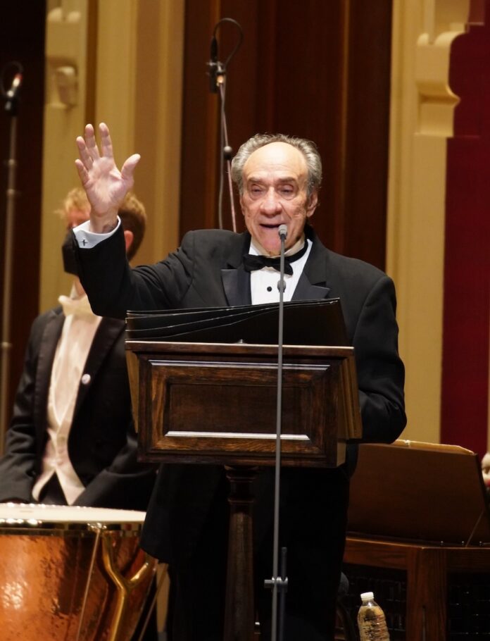 F. Murray Abraham performing behind a podium onstage with the Pittsburgh Symphony Orchestra.