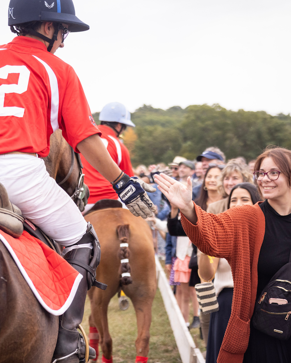A woman in a rust colored sweater shaking the hand of a polo player in a red shirt on a horse.
