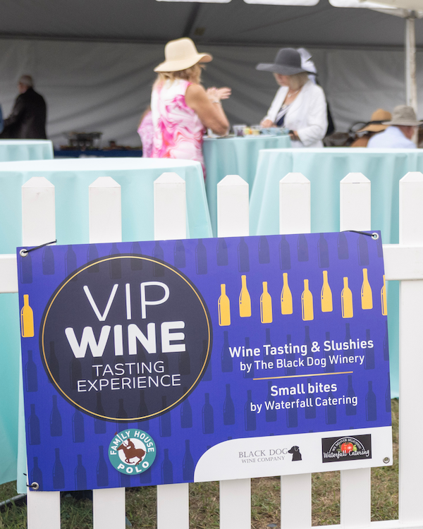 A photo of a sign hanging on a white picket fence that advertises the VIP wine tasting experience at the Family House Polo Match.