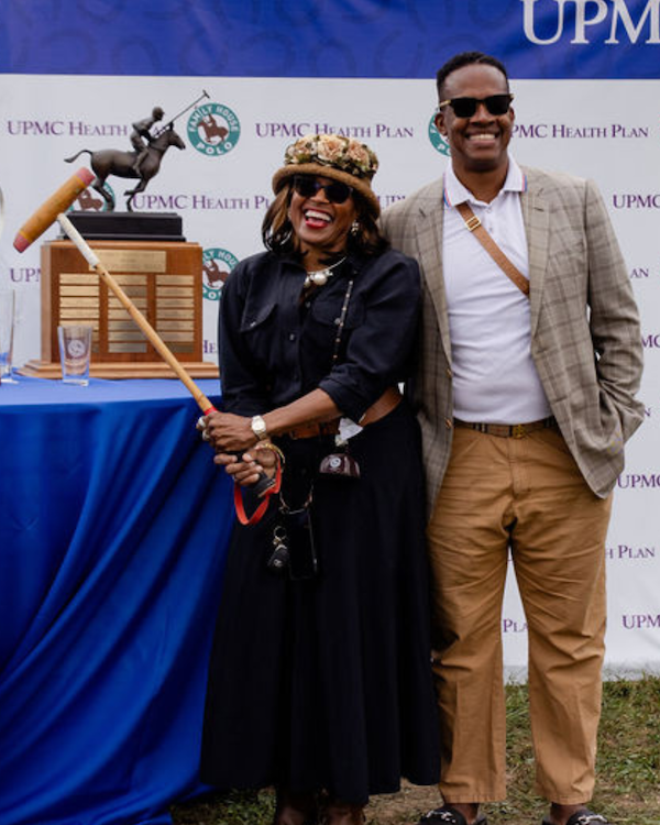A couple standing in front of the Family House Polo trophy and the woman is holding a polo mallet.