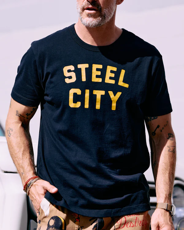 A man in a black shirt with yellow font that says "steel city" on it.