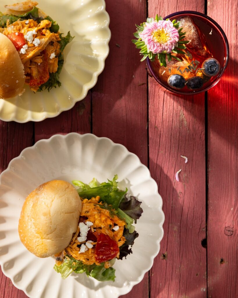 A white plate with a chicken sandwich on top with toppings and another plate of a slider above that and a glass of tea in the upper right hand corner.