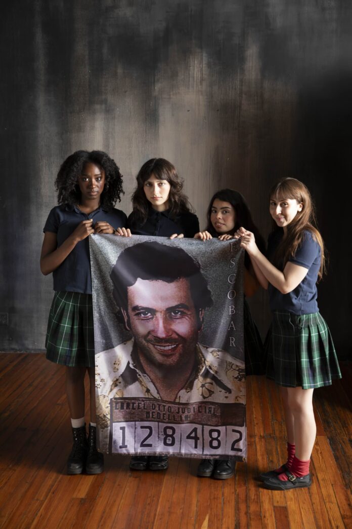 Four girls hold up a picture of a drug lord as they stand in school girl outfits.