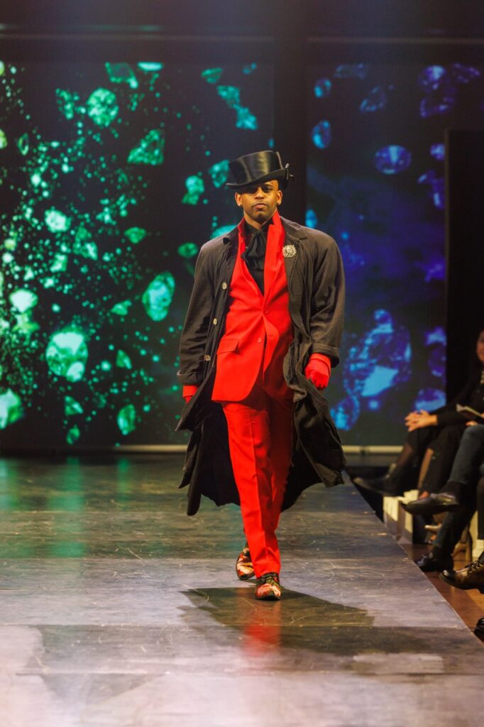 A man walking down a runway in a red suit with a black top hat and black jacket over top.